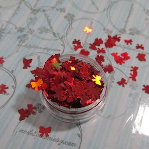 May include: A small jar filled with red glitter shaped like bows. The glitter is iridescent and has a holographic finish. The jar is on a blue and white patterned surface.