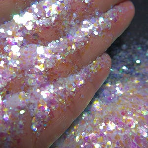 May include: Close-up of iridescent glitter in various shapes and sizes. The glitter is a mix of pink, white, and yellow, with a sparkling, reflective quality. Some glitter is on a surface, and some is on a hand.