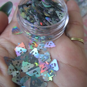 Op de afbeelding: Een klein, doorzichtig potje gevuld met regenboogkleurig, holografisch glitter in de vorm van geesten. De glitter is verspreid over een hand.