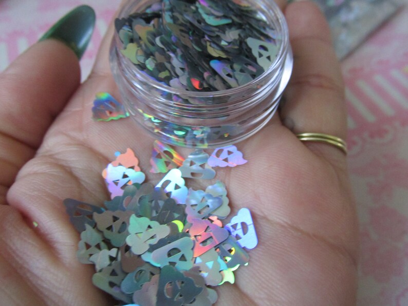 Op de afbeelding: Een klein, doorzichtig potje gevuld met regenboogkleurig, holografisch glitter in de vorm van geesten. De glitter is verspreid over een hand.