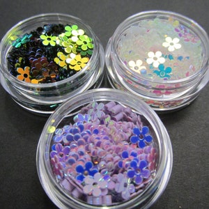 May include: Three small jars filled with iridescent flower-shaped glitter. The jars are clear plastic and the glitter is in shades of green, purple, and white.