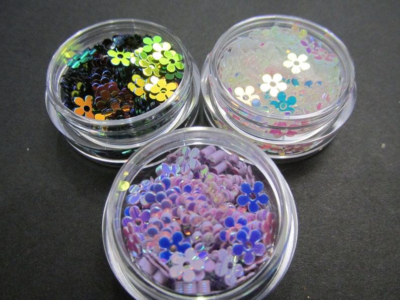 May include: Three small jars filled with iridescent flower-shaped glitter. The jars are clear plastic and the glitter is in shades of green, purple, and white.