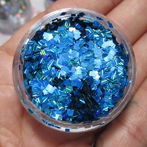 May include: A small, clear plastic pot filled with bright blue glitter. The glitter consists of square-shaped pieces that shimmer and reflect light. The pot is held in a hand, highlighting the glitter's texture and colour.