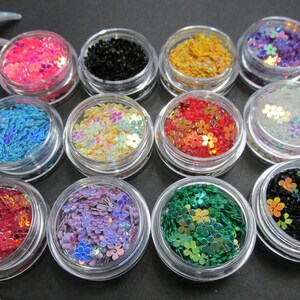 May include: Twelve small jars filled with colorful iridescent flower-shaped glitter. The glitter is in various colors, including pink, blue, green, red, gold, and black.