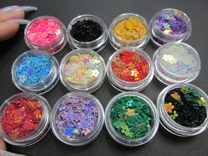 May include: Twelve small jars filled with colorful iridescent flower-shaped glitter. The glitter is in various colors, including pink, blue, green, red, gold, and black.