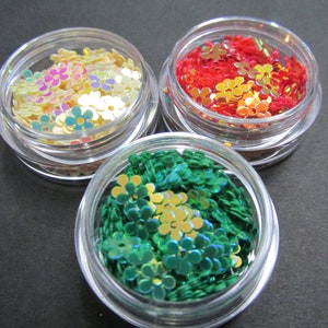 May include: Three small clear jars filled with iridescent flower-shaped glitter. The jars contain green, red, and multi-colored glitter.