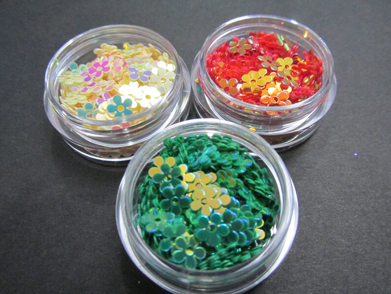May include: Three small clear jars filled with iridescent flower-shaped glitter. The jars contain green, red, and multi-colored glitter.