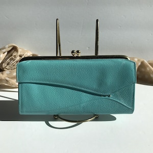 May include: A turquoise leather clutch with a gold clasp and frame. The rectangular wallet has a curved flap and is displayed on a gold metal stand. The clutch is a solid color and has a textured surface.