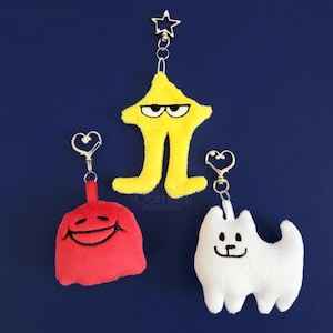 May include: Three plush keyrings on a blue background. One is a yellow star with black eyes and legs, another is a red smiling face, and the third is a white dog with a black face. Each keyring has a metal clasp.