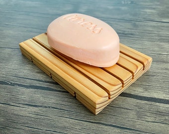 May include: A wooden soap dish with a light brown finish and a pink bar of soap on top. The soap has the word "Cleans" embossed on it.