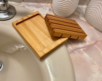 May include: Two wooden soap dishes, one with a raised design and the other flat, are sitting on a white porcelain sink.