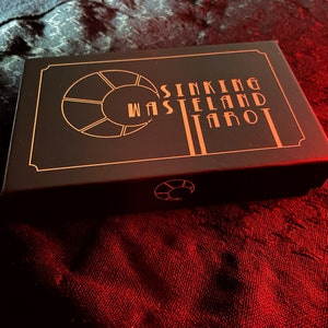 May include: A black tarot card box with gold lettering and design. The text on the box reads "Sinking Wasteland Tarot". The box is rectangular and sits on a red and black textured surface.