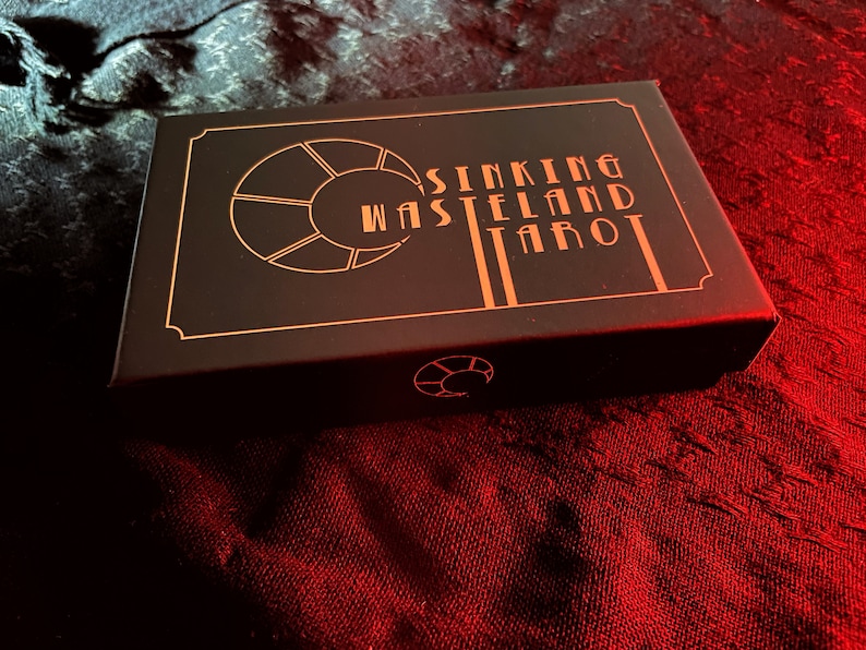 May include: A black tarot card box with gold lettering and design. The text on the box reads "Sinking Wasteland Tarot". The box is rectangular and sits on a red and black textured surface.