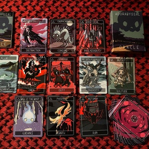 May include: A set of 14 tarot cards with black backgrounds and colorful illustrations of mythical creatures. The cards are laid out on a red and white checkered surface. The cards are part of the "Adorabyssal Oracle" deck.
