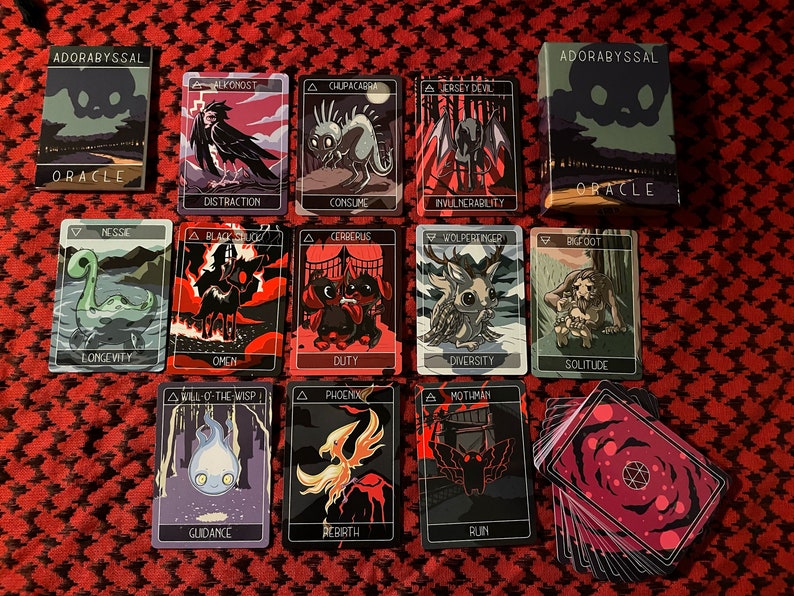 May include: A set of 14 tarot cards with black backgrounds and colorful illustrations of mythical creatures. The cards are laid out on a red and white checkered surface. The cards are part of the "Adorabyssal Oracle" deck.