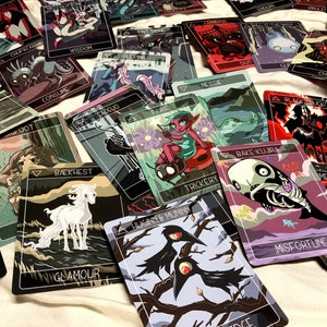 May include: A set of playing cards with colorful illustrations and text. The cards feature a variety of characters and creatures, including a white unicorn, a black bird, a skull, and a green elf. The cards are designed in a whimsical style with a dark background.