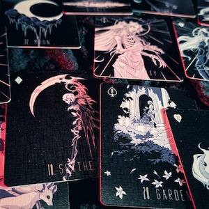 May include: A collection of tarot cards with dark backgrounds and illustrations in shades of pink, white, and blue. The cards feature images of animals, figures, and celestial bodies. The cards have red edges.