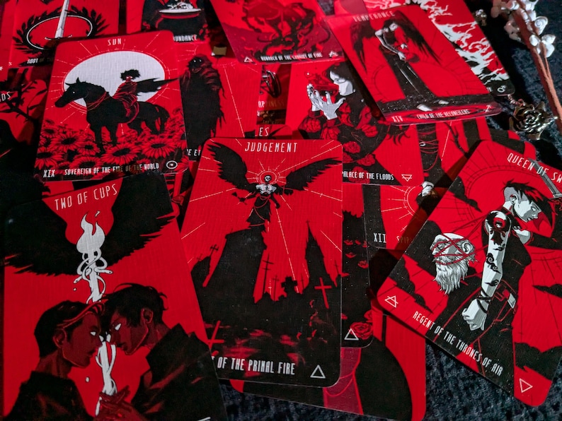 May include: A set of tarot cards with red and black designs. The cards feature illustrations of angels, cups, and other symbolic imagery. The cards are arranged in a spread, with the text "JUDGEMENT", "TWO OF CUPS", "QUEEN OF SW", "REGENT OF THE THRONES OF AIR", and "RACE OF THE FLOODS" visible.