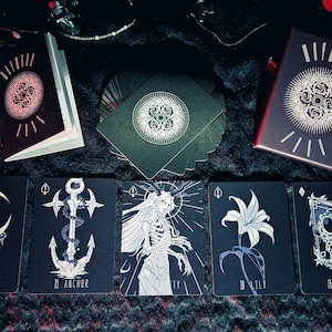 May include: A deck of tarot cards with a dark aesthetic. The cards feature illustrations of a crescent moon, an anchor, a skeletal figure, a lily, and a book. The card backs and box have a circular design. The color palette is black, white, and red.