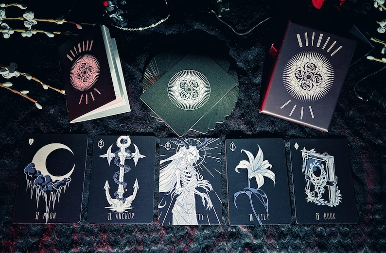 May include: A deck of tarot cards with a dark aesthetic. The cards feature illustrations of a crescent moon, an anchor, a skeletal figure, a lily, and a book. The card backs and box have a circular design. The color palette is black, white, and red.