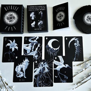 May include: A black tarot card deck with illustrated cards featuring a gothic theme. The deck includes a box, a booklet, and a stack of cards. The cards feature illustrations of figures, animals, and celestial bodies in white against a black background.
