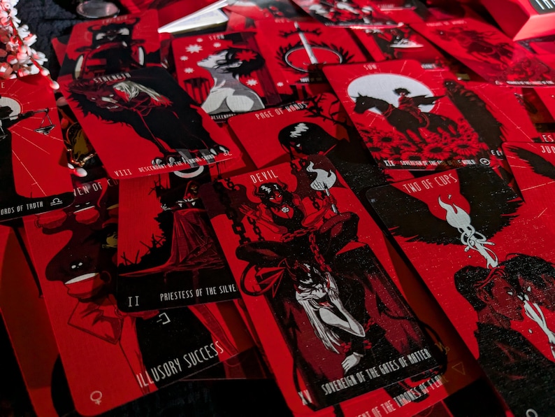 May include: A set of tarot cards with red and black illustrations. The cards feature various characters and symbols, including a devil, a priestess, and a sovereign. The cards are laid out on a black surface.