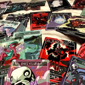 May include: A collection of colorful playing cards with illustrations of various mythical creatures, including a black shuck, a bake-kujira, a dullahan, a cerrechung, a will-o'-the-wisp, a demon, a nessie, a trickster, a fairie, a sleeping beauty, a cait sith, a cadejo, and an endure. The cards are laid out on a white surface.