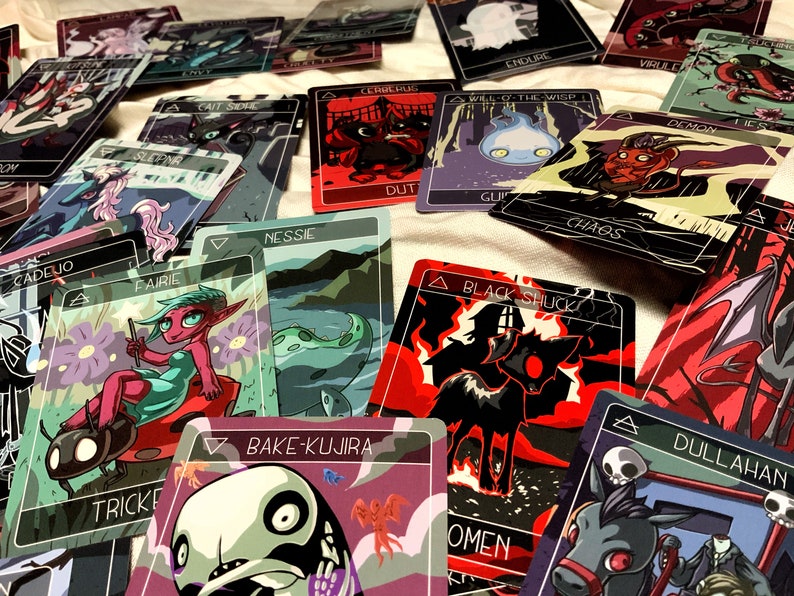 May include: A collection of colorful playing cards with illustrations of various mythical creatures, including a black shuck, a bake-kujira, a dullahan, a cerrechung, a will-o'-the-wisp, a demon, a nessie, a trickster, a fairie, a sleeping beauty, a cait sith, a cadejo, and an endure. The cards are laid out on a white surface.