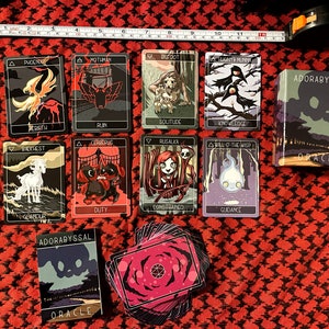 May include: A set of tarot cards with black and white illustrations of various creatures and symbols. The cards are laid out on a red and black patterned surface. The cards are part of the "Adorabyssal Oracle" deck.