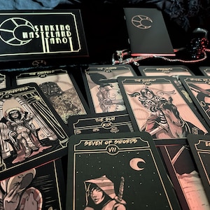 May include: A tarot card deck with the title "Sinking Wasteland Tarot" on the box. The cards feature illustrations in black, gold, and red tones. Several cards are spread out, displaying various tarot imagery, including "The High Priestess" and "Seven of Swords".