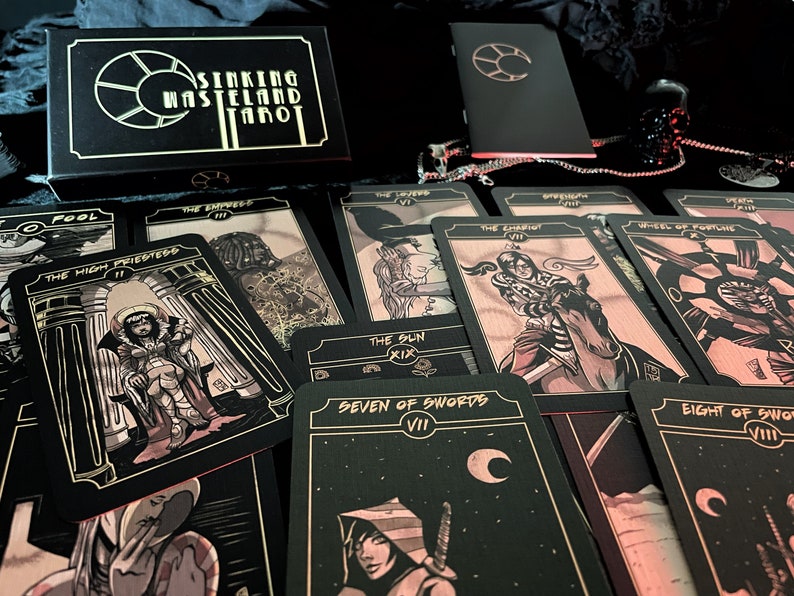 May include: A tarot card deck with the title "Sinking Wasteland Tarot" on the box. The cards feature illustrations in black, gold, and red tones. Several cards are spread out, displaying various tarot imagery, including "The High Priestess" and "Seven of Swords".
