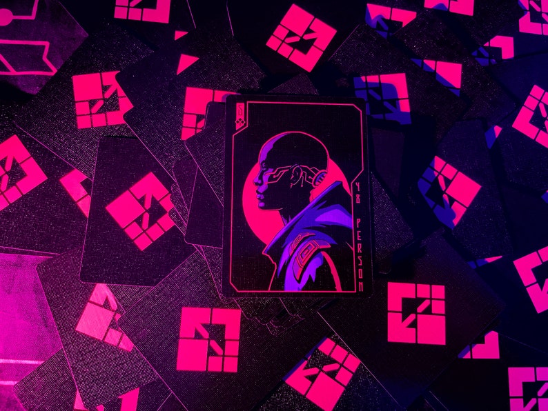 K&ouml;nnte beinhalten: Schwarze Spielkarten mit einem neonrosa geometrischen Design und einer zentralen Karte mit dem Profil einer Person mit futuristischen technischen Details. Die Karte tr&auml;gt den Text "YR PERSON". Der Hintergrund ist dunkel.