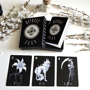 May include: A black tarot card deck with a floral design on the box and individual cards. The cards feature illustrations of a lily, a wolf, and a scythe. The deck is displayed with white floral branches and a black skull.