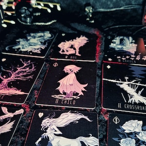 May include: A tarot card deck with a black and red box and several cards laid out. The cards feature illustrations of a tree, a child, a wolf, and other mystical images. The deck is designed with a dark, gothic aesthetic.