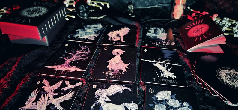 May include: A tarot card deck with a black and red box and several cards laid out. The cards feature illustrations of a tree, a child, a wolf, and other mystical images. The deck is designed with a dark, gothic aesthetic.