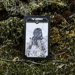 May include: A black and white tarot card featuring an illustration of a woman with star-adorned dreadlocks, surrounded by vines. The card reads "THE EMPRESS" at the top, with the Roman numeral "III" below. The card is set against a mossy background.