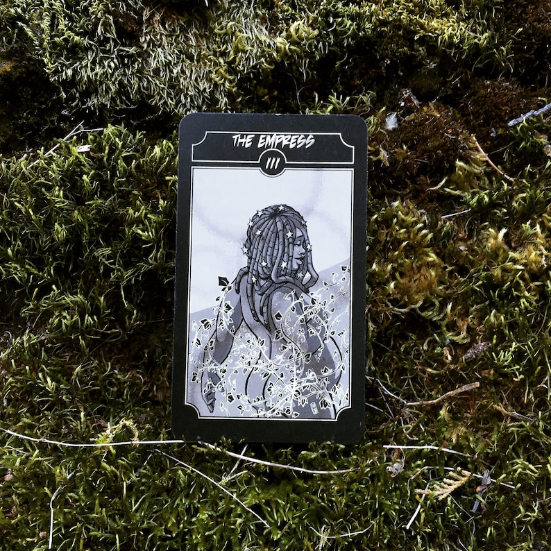 May include: A black and white tarot card featuring an illustration of a woman with star-adorned dreadlocks, surrounded by vines. The card reads "THE EMPRESS" at the top, with the Roman numeral "III" below. The card is set against a mossy background.