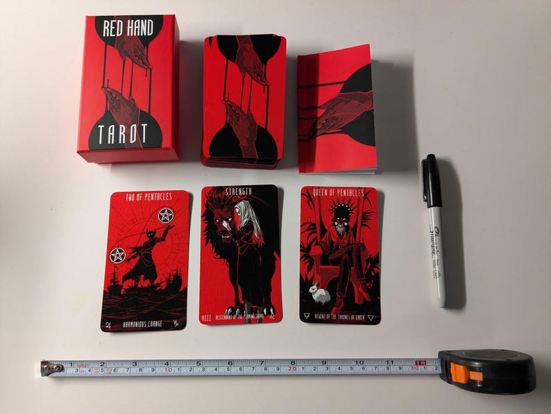 May include: Red Hand Tarot deck with a red box and three cards showing Strength, Queen of Pentacles, and Two of Pentacles. The cards feature black and white illustrations on a red background.
