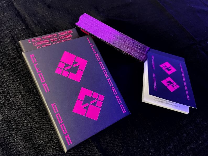 K&ouml;nnte beinhalten: Ein Retro-Futuristic-Cyberpunk-Lenormand-Deck mit 59 einzigartigen Illustrationen. Das Deck und die Box sind dunkelblau mit leuchtend rosa geometrischen Designs und Text. Das Deck ist teilweise aufgef&auml;chert und zeigt die Kartenkanten.