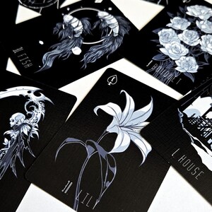 May include: A collection of black playing cards with white and gray illustrations. The cards feature images of a wolf, fish, roses, a scythe, a lily, and a house. Each card has a unique design and number.