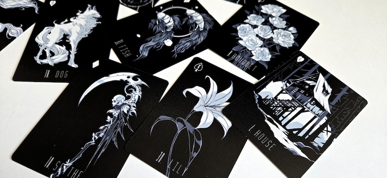 May include: A collection of black playing cards with white and gray illustrations. The cards feature images of a wolf, fish, roses, a scythe, a lily, and a house. Each card has a unique design and number.
