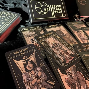 May include: A tarot card deck with black cards and gold illustrations, featuring cards like "The Fool" and "King of Cups." The deck is in a black box with the words "Sinking Wasteland Tarot." The edges of the cards are red.