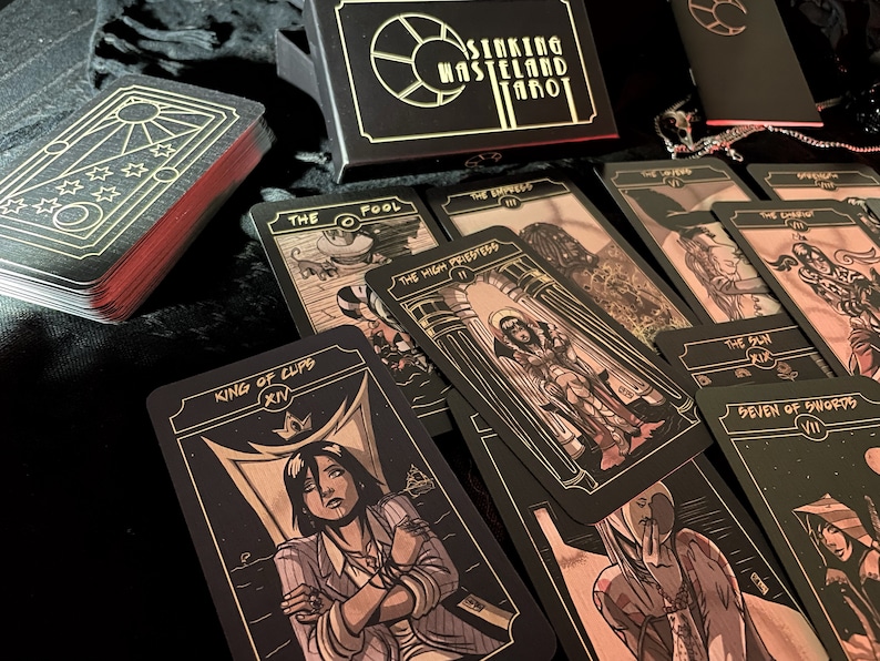 May include: A tarot card deck with black cards and gold illustrations, featuring cards like "The Fool" and "King of Cups." The deck is in a black box with the words "Sinking Wasteland Tarot." The edges of the cards are red.
