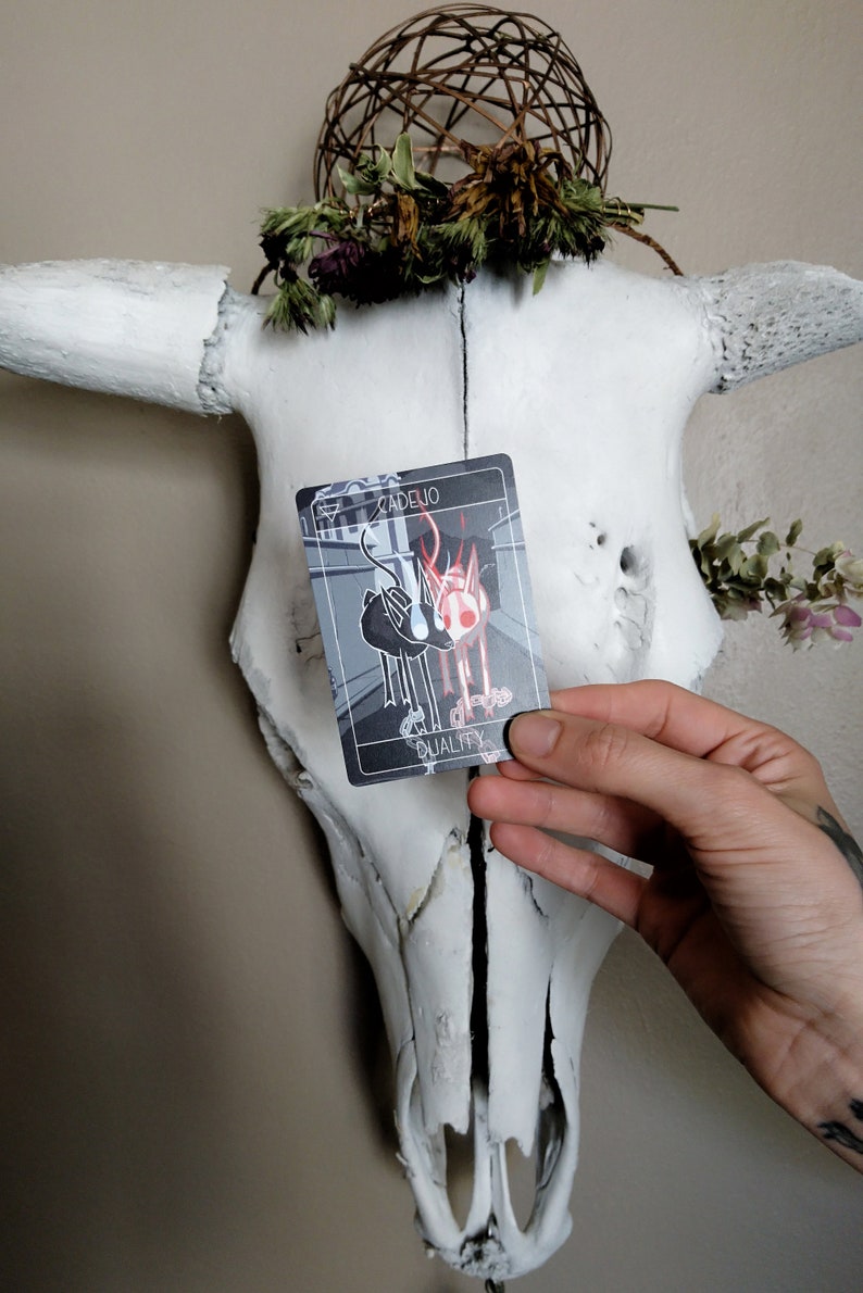 May include: A black and white tarot card with a red and white illustration of two animals. The card is labeled "Cadelio" and "Duality". The card is held in front of a white animal skull with a wreath of dried flowers.