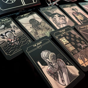 May include: A tarot card deck with black cards and gold accents. The cards feature detailed illustrations in a comic book style. The cards are laid out on a dark surface, with the box visible in the background. The cards include "The Fool" and "The Moon".