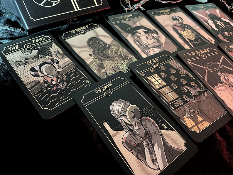 May include: A tarot card deck with black cards and gold accents. The cards feature detailed illustrations in a comic book style. The cards are laid out on a dark surface, with the box visible in the background. The cards include "The Fool" and "The Moon".