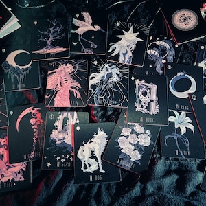May include: A collection of tarot cards with dark, gothic-inspired illustrations. The cards feature images of a wolf, a crescent moon, a skeleton, and other mystical symbols in shades of black, white, and pink. The cards are scattered on a dark surface.