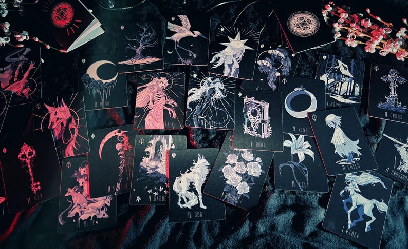 May include: A collection of tarot cards with dark, gothic-inspired illustrations. The cards feature images of a wolf, a crescent moon, a skeleton, and other mystical symbols in shades of black, white, and pink. The cards are scattered on a dark surface.