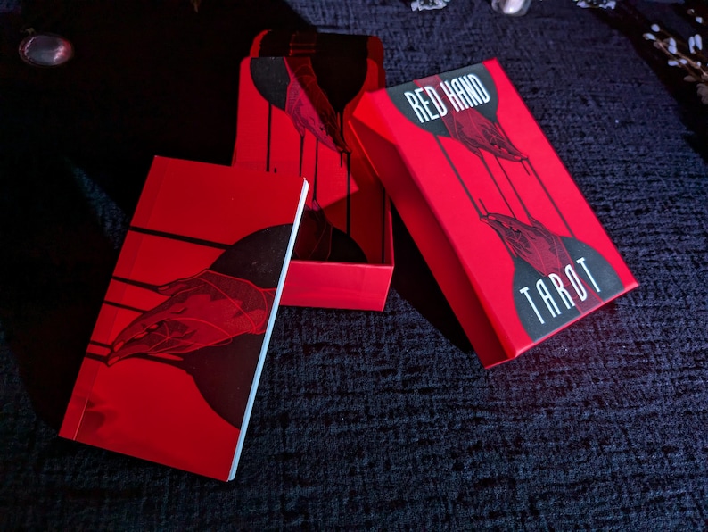 May include: Red Hand Tarot deck box with a red background and black text. The box is open and the cards are visible inside. The box has a graphic of two hands with red and black details.