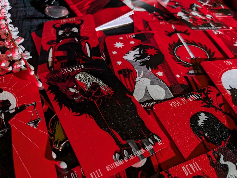 May include: A set of tarot cards with red backgrounds and black illustrations. The cards feature various symbols and characters, including a lion, a sword, and a star. The cards are labeled with their corresponding names, such as "Strength", "Page of Wands", and "Sun".
